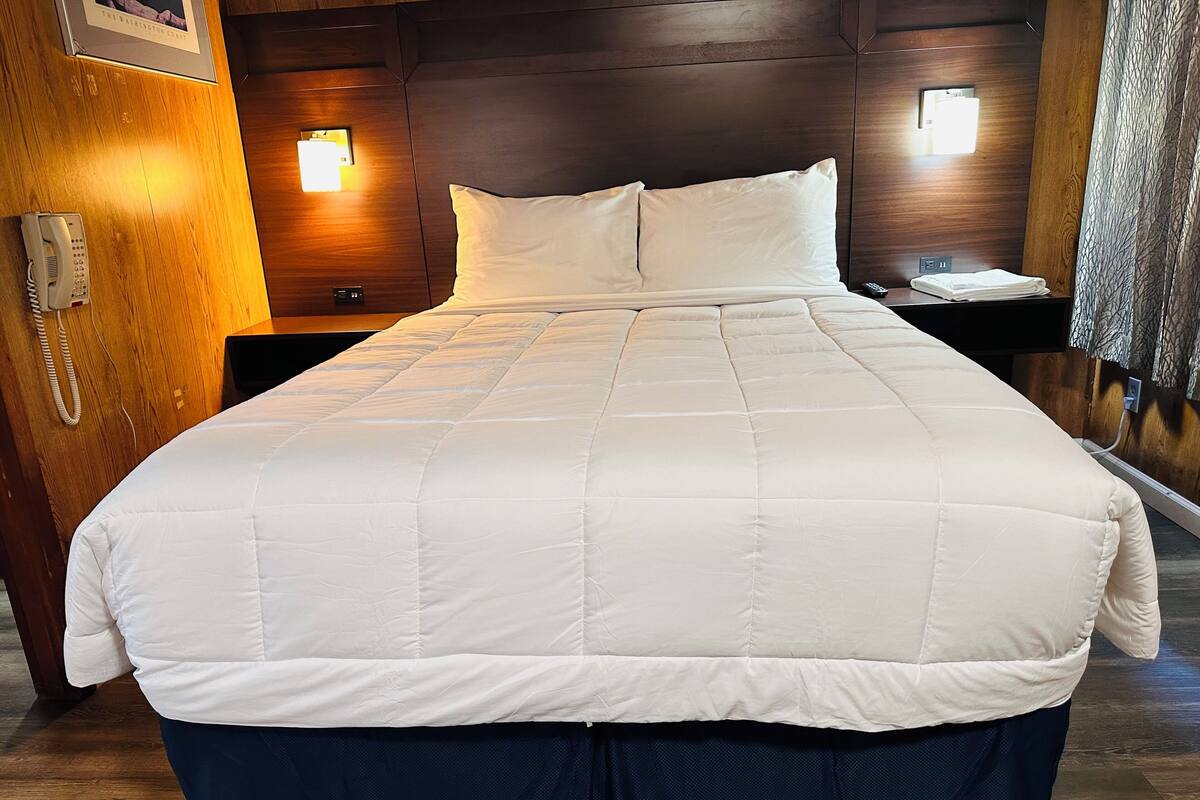 Un letto rifatto con copriletto bianco e gonna a balze blu scuro, una testiera in legno, un comodino con una lampada e un telefono a muro.