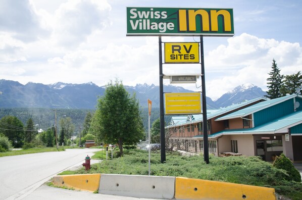 A Swiss Village Inn sign with an RV sites sign below it, set against a backdrop of mountains and a building with a green roof.