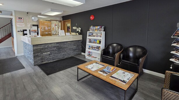 A reception area with a reception desk, a clock, a red emergency light, a white bookshelf with magazines, two leather chairs, and a wooden coffee table with brochures. - Swiss Village Inn (Golden)