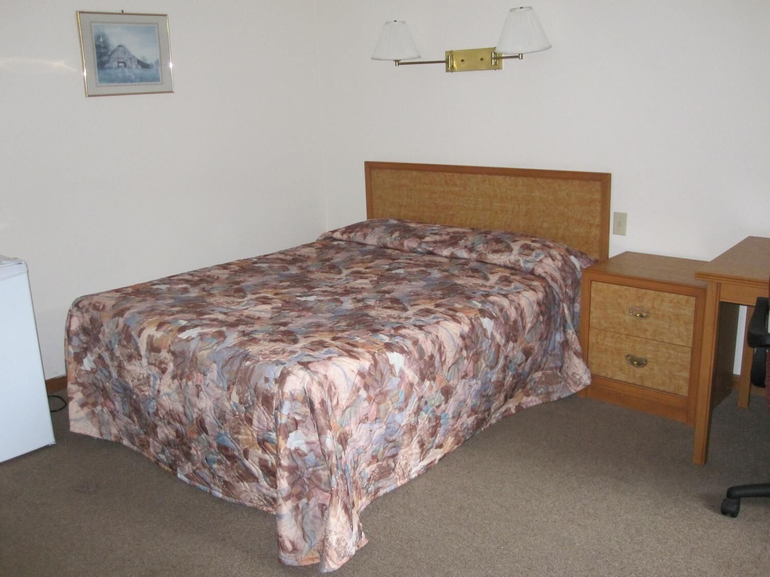 A bedroom with a bed, a nightstand, a desk, and a wall-mounted lamp.