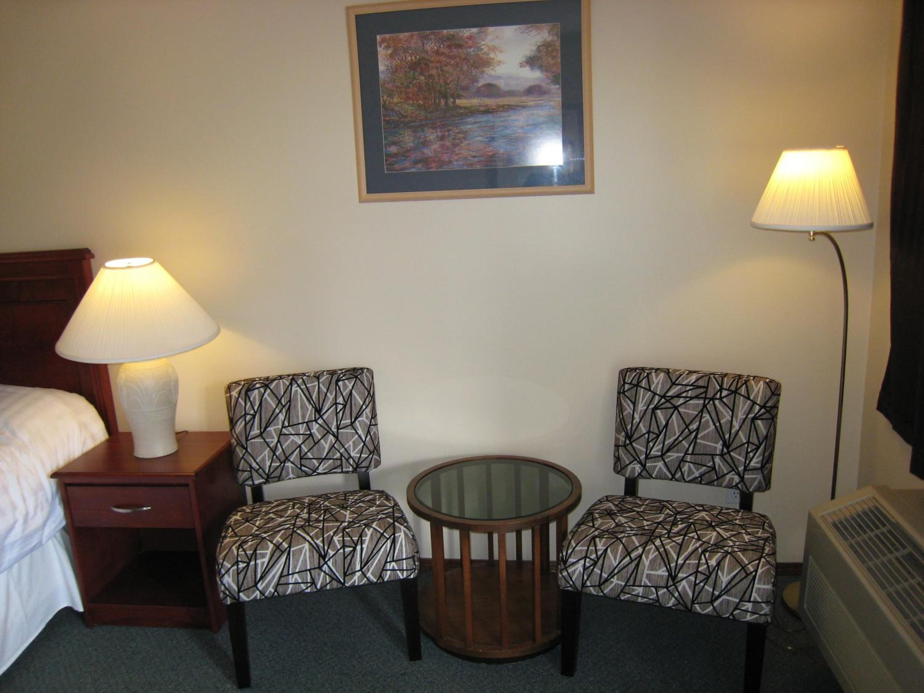 A hotel room with a bed, two patterned chairs, a small round table, a nightstand with a lamp, a framed picture on the wall, and a standing lamp.