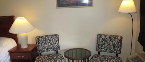 A hotel room with a bed, two patterned chairs, a small round table, a nightstand with a lamp, a framed picture on the wall, and a standing lamp.