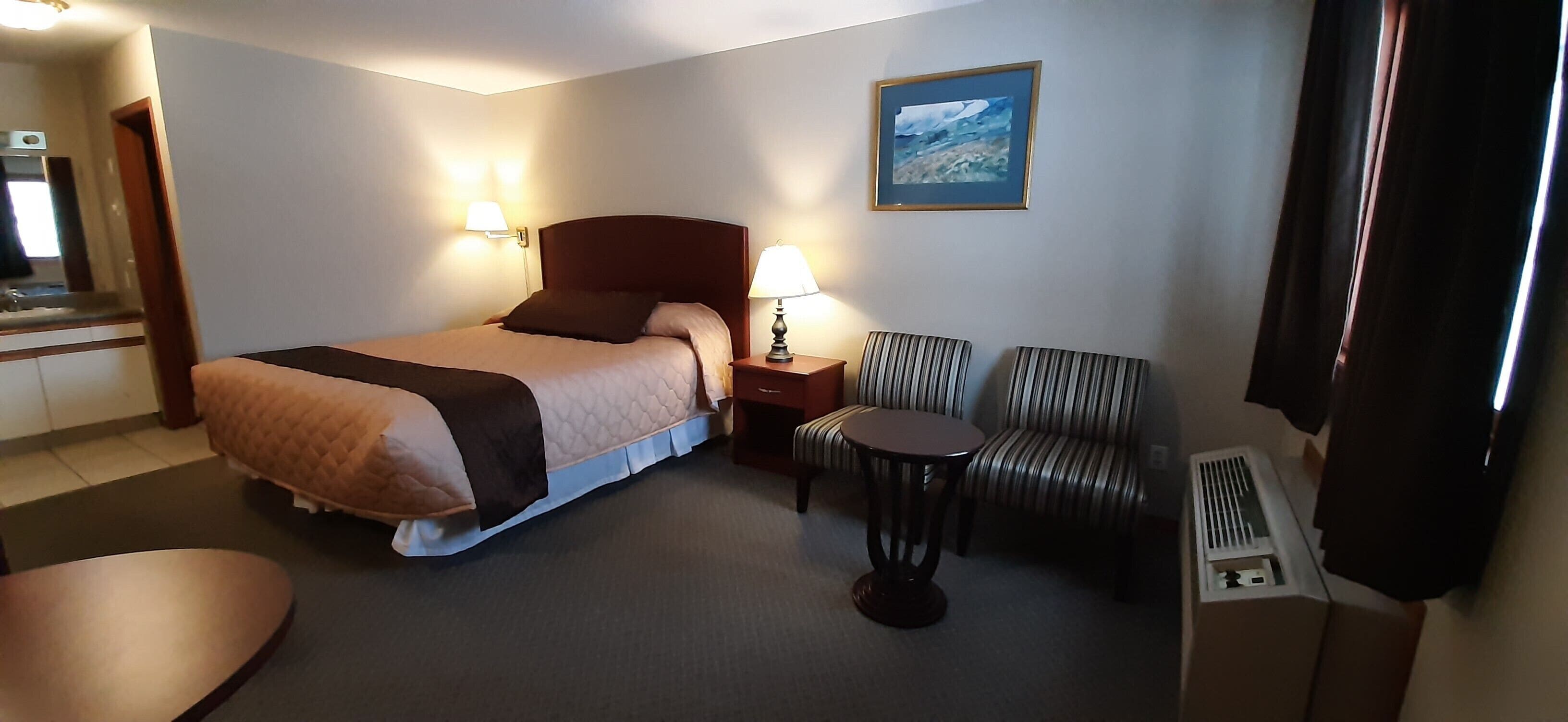 Quarto de hotel com cama, mesa redonda pequena, duas cadeiras listradas, mesa de cabeceira com abajur, quadro na parede e janela com cortinas.