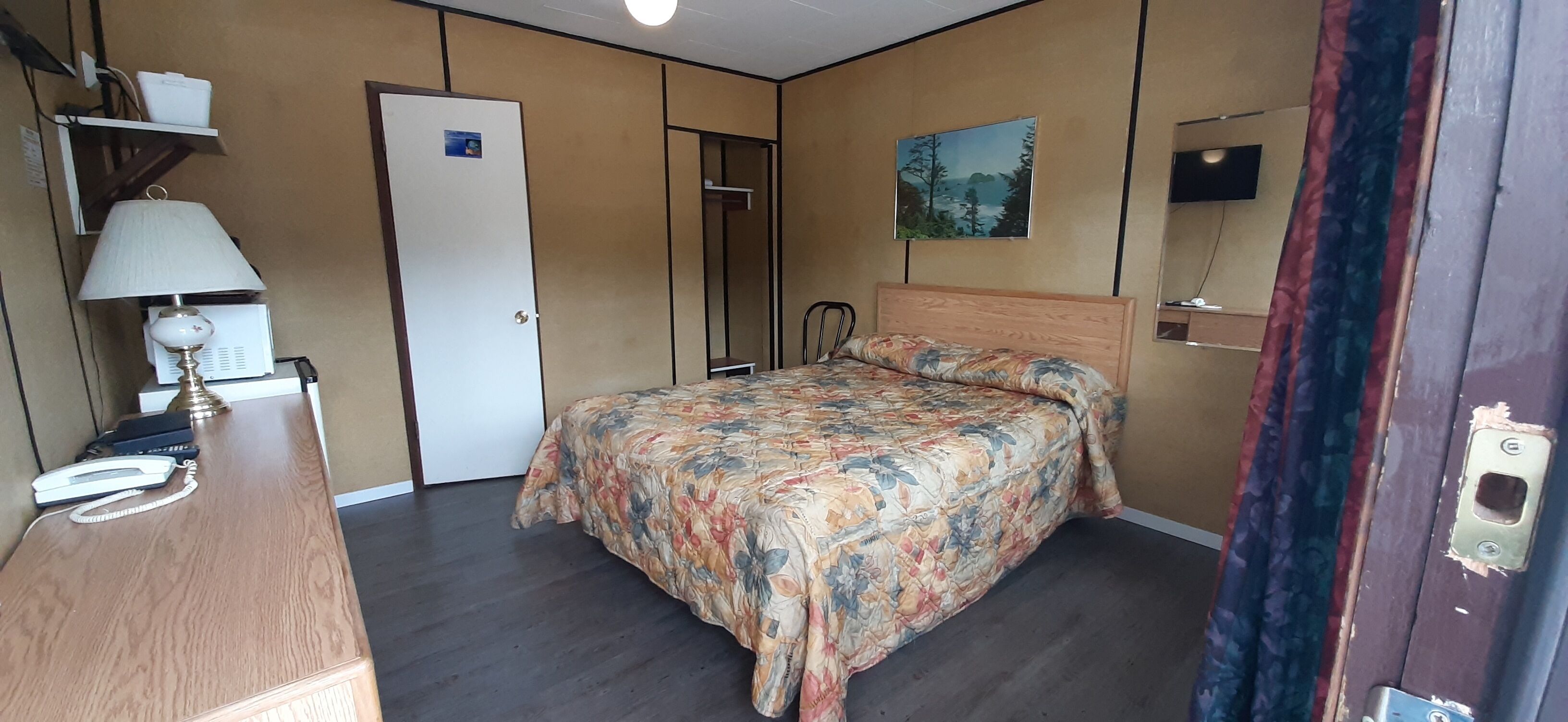 Ein Schlafzimmer mit Bett, Schreibtisch mit Lampe, einer Tür und einem Bild an der Wand.