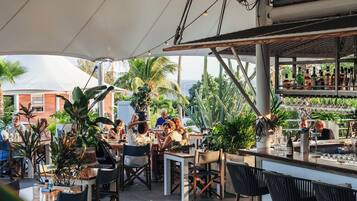 Un área de comedor al aire libre con mesas y sillas, una barra con botellas y vista al océano.