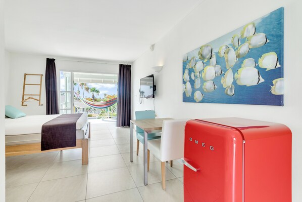 A modern hotel room with a bed, a dining table, chairs, a red refrigerator, a hammock, and a large painting of fish.