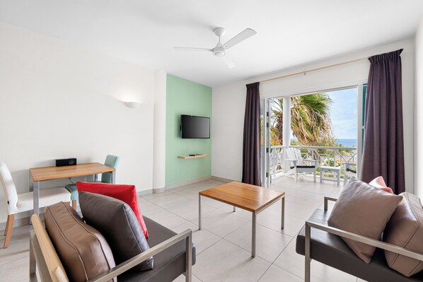 A modern living room with a sofa, a coffee table, a TV mounted on the wall, and a balcony with a view of the sea. - Chogogo Dive & Beach Resort Curacao (Jan Thiel)