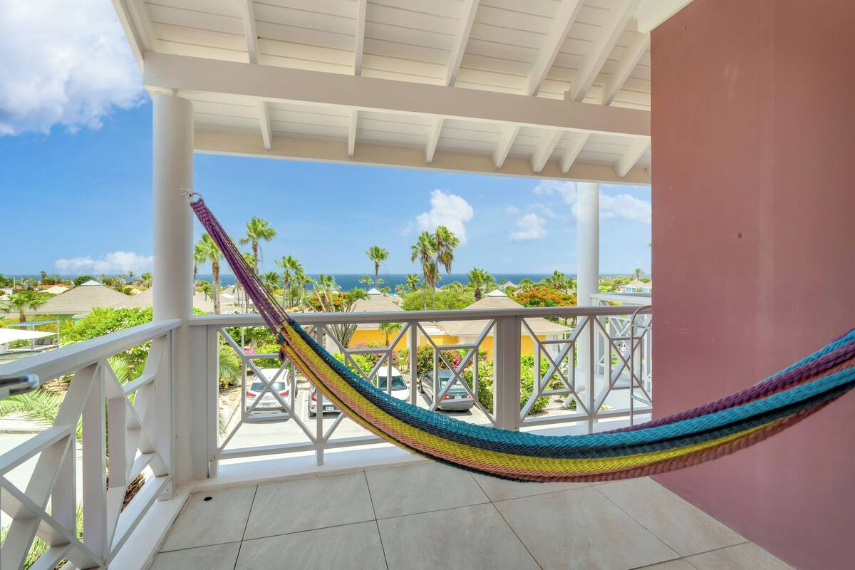 A balcony with a hammock, white railings, and a view of the ocean and palm trees.