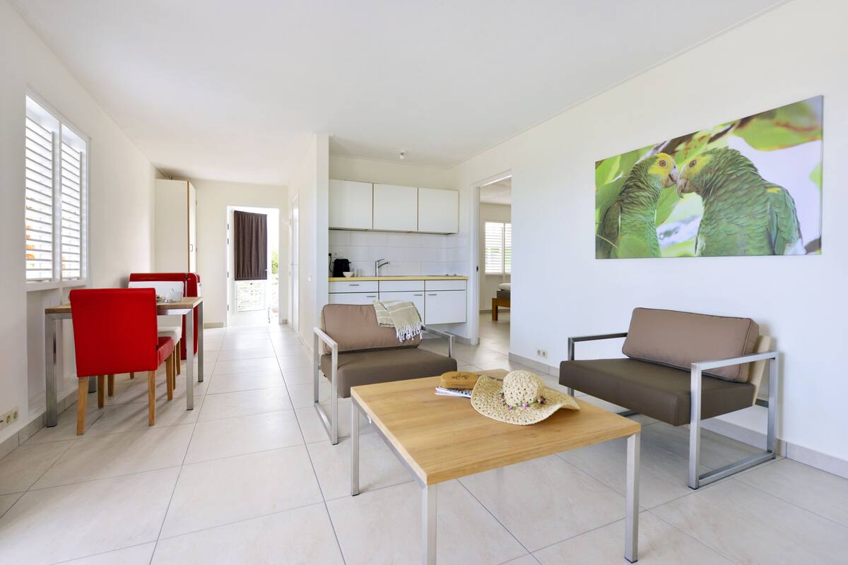 A modern living room with a wooden coffee table, red chairs, and a sofa. A kitchen is visible in the background.