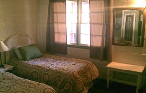 A hotel room with two beds, a nightstand, a chair, a small table, and a window with curtains.