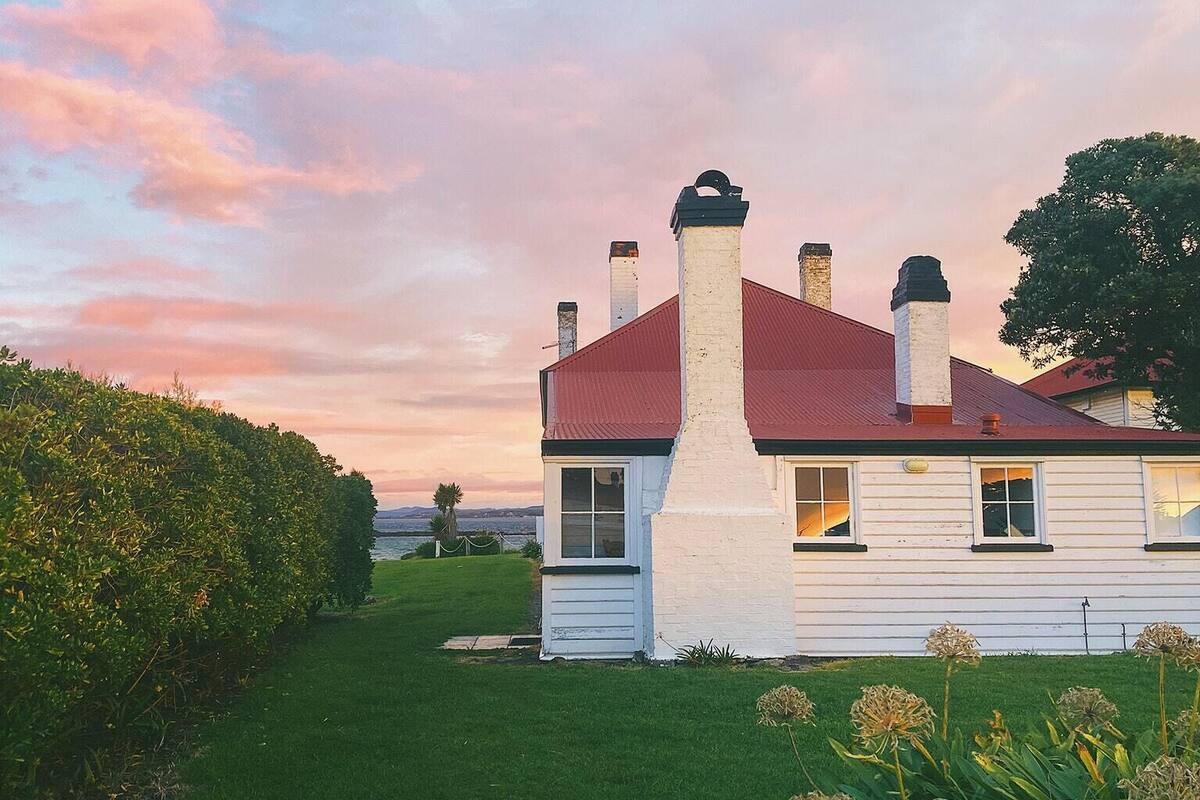 Una casa blanca con techo rojo y varias chimeneas, rodeada de un césped cuidado y plantas con flores.