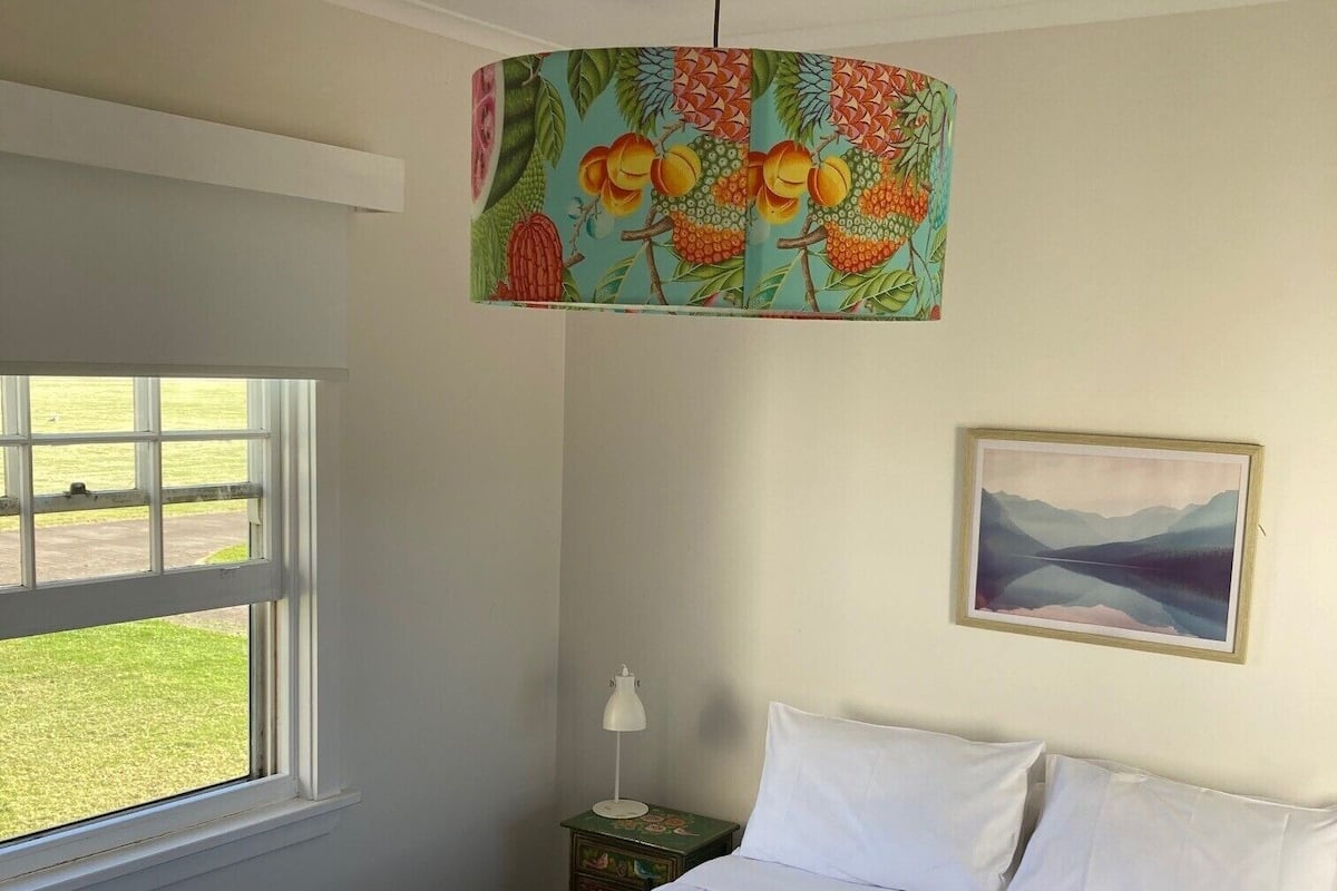 Un dormitorio con cama, ventana, mesita de noche, lámpara, un cuadro y una lámpara colgante de colores.
