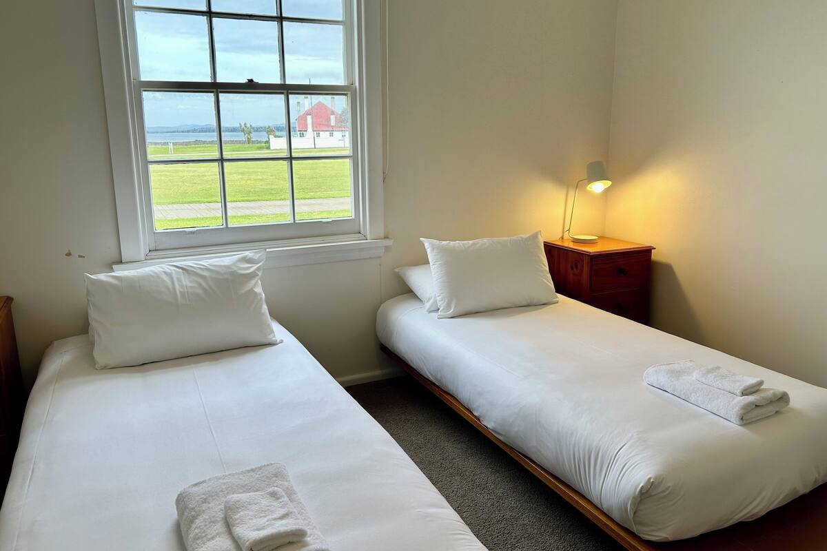 Una habitación con dos camas individuales, una ventana que ofrece vista a un edificio rojo y una zona ajardinada, una mesita de noche con lámpara y suelo alfombrado.