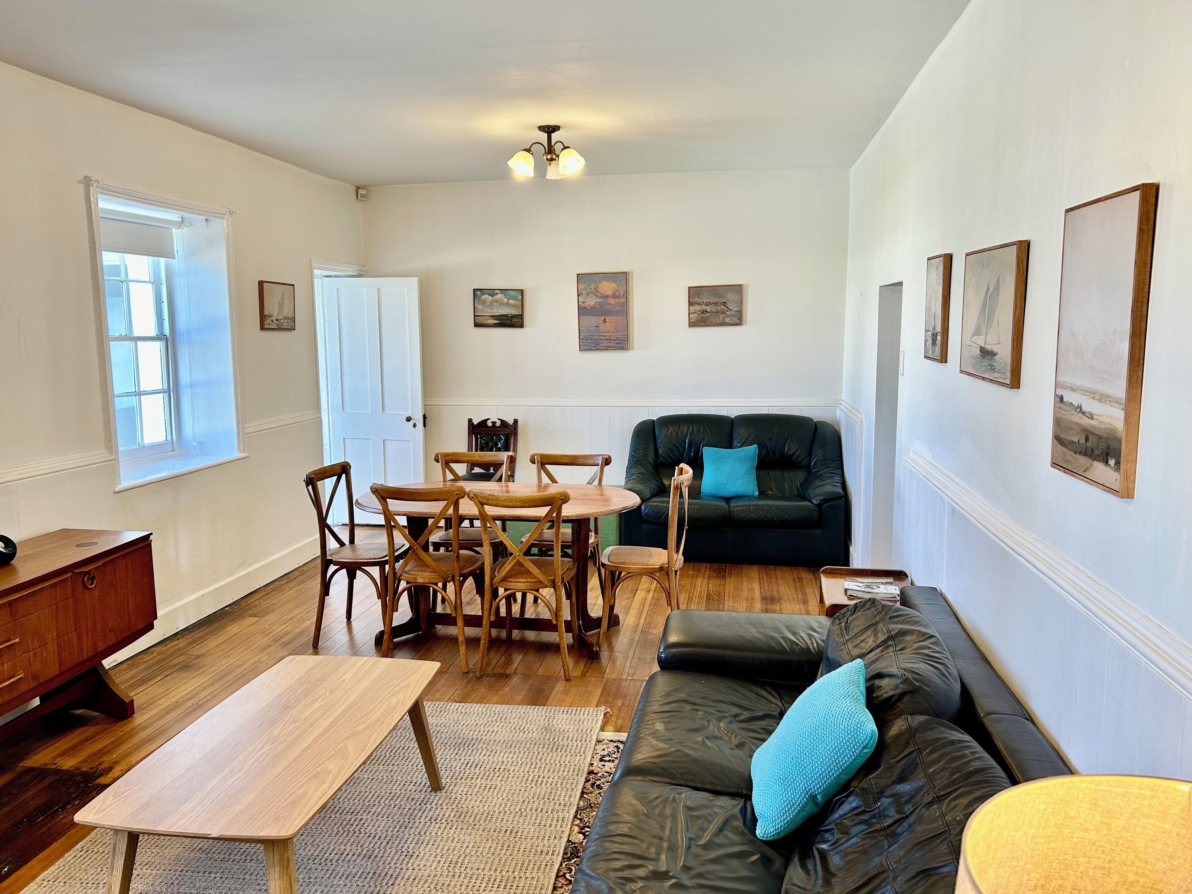 A living room with a dining table, chairs, a sofa, and a coffee table. There are framed pictures on the wall and a door leading to another room.