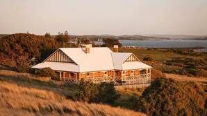 Una casa grande con techo blanco y varias chimeneas, rodeada de hierba alta y árboles, con vista a un cuerpo de agua.