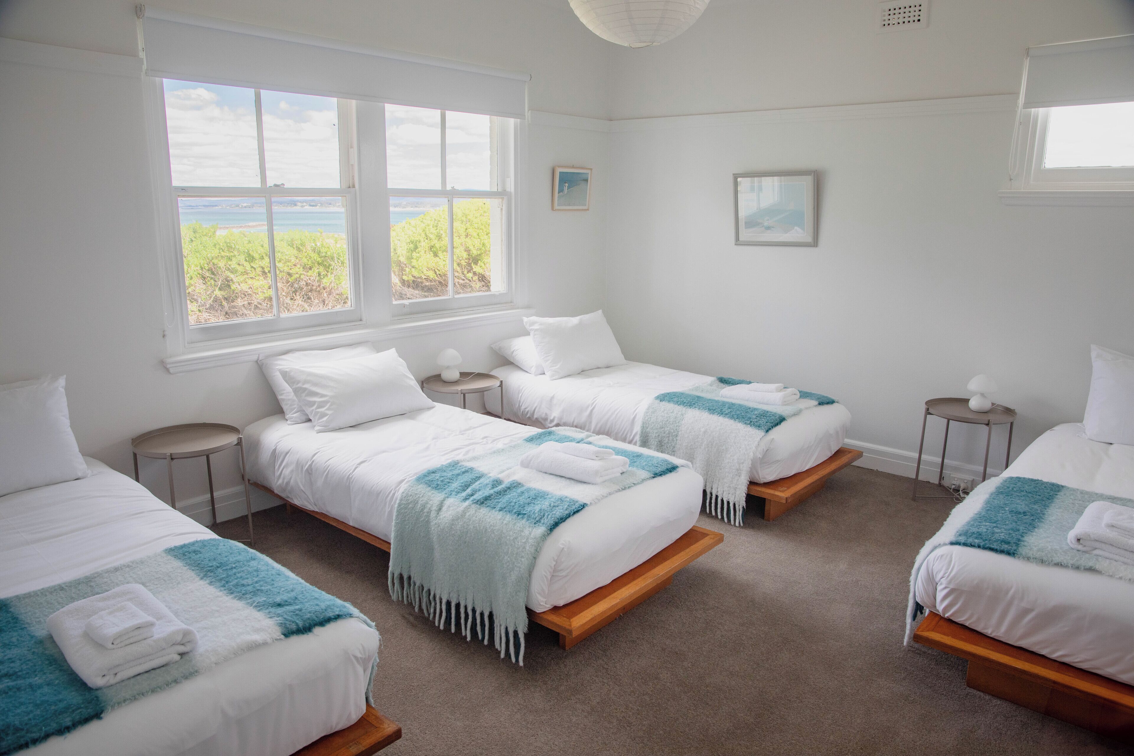 Une chambre d’hôtel avec trois lits, une grande fenêtre donnant sur la mer et une petite table ronde avec une lampe.