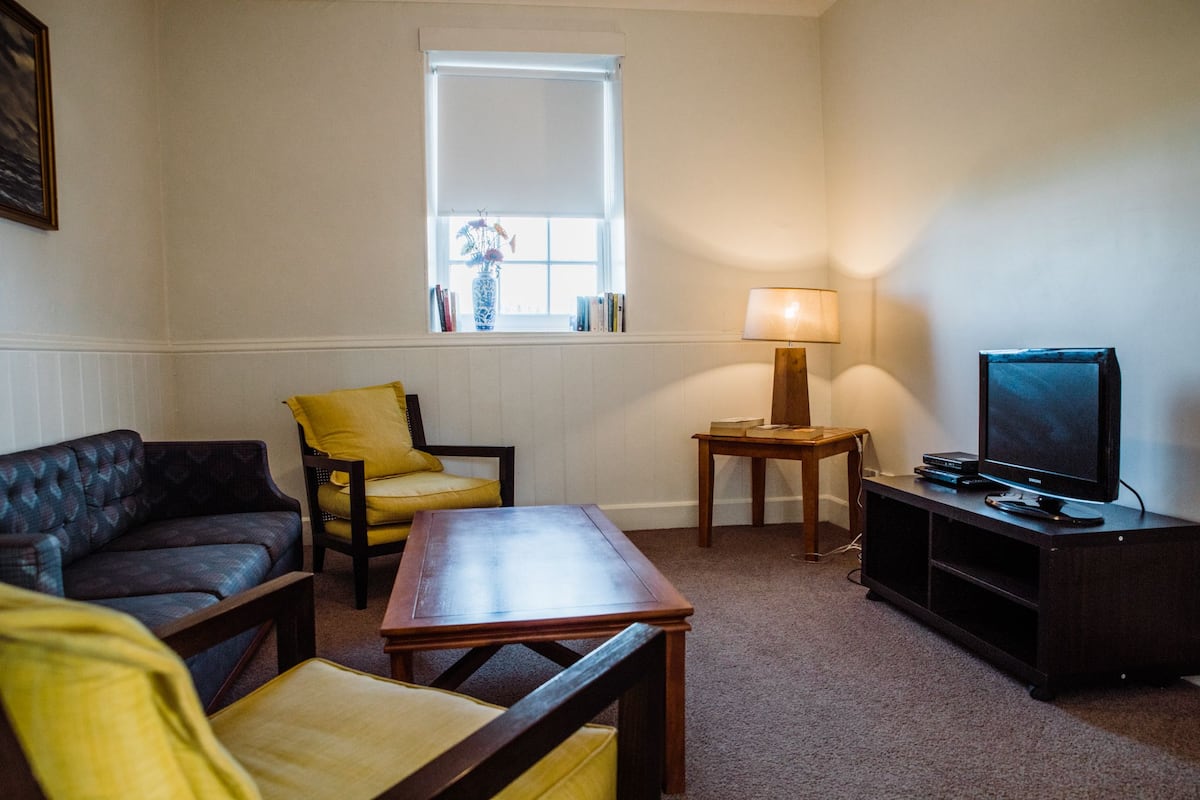 Una sala de estar con sofá, sillón, mesa, lámpara, televisión y una ventana con una planta.