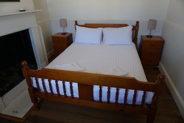A neatly made bed with white linens and pillows, flanked by two bedside tables with lamps, in a room with wooden flooring and a fireplace.
