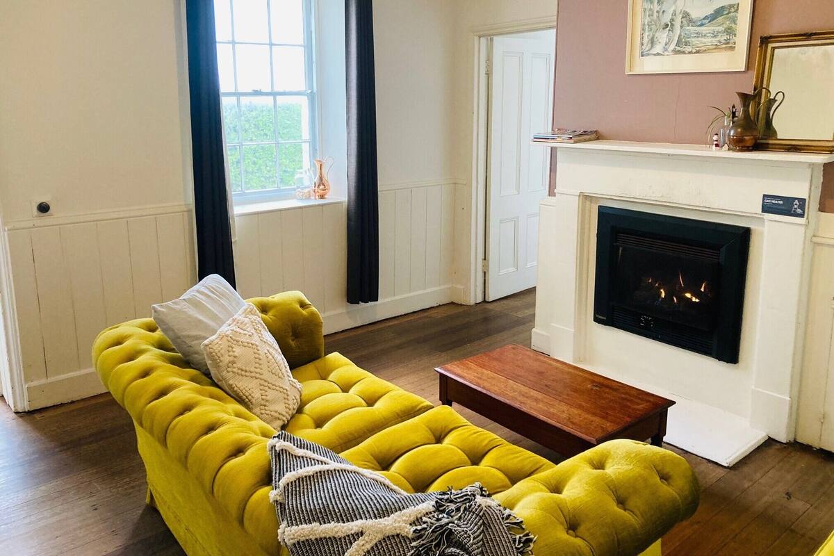 Una sala de estar con un sofá amarillo, una chimenea y un ventanal con cortinas.