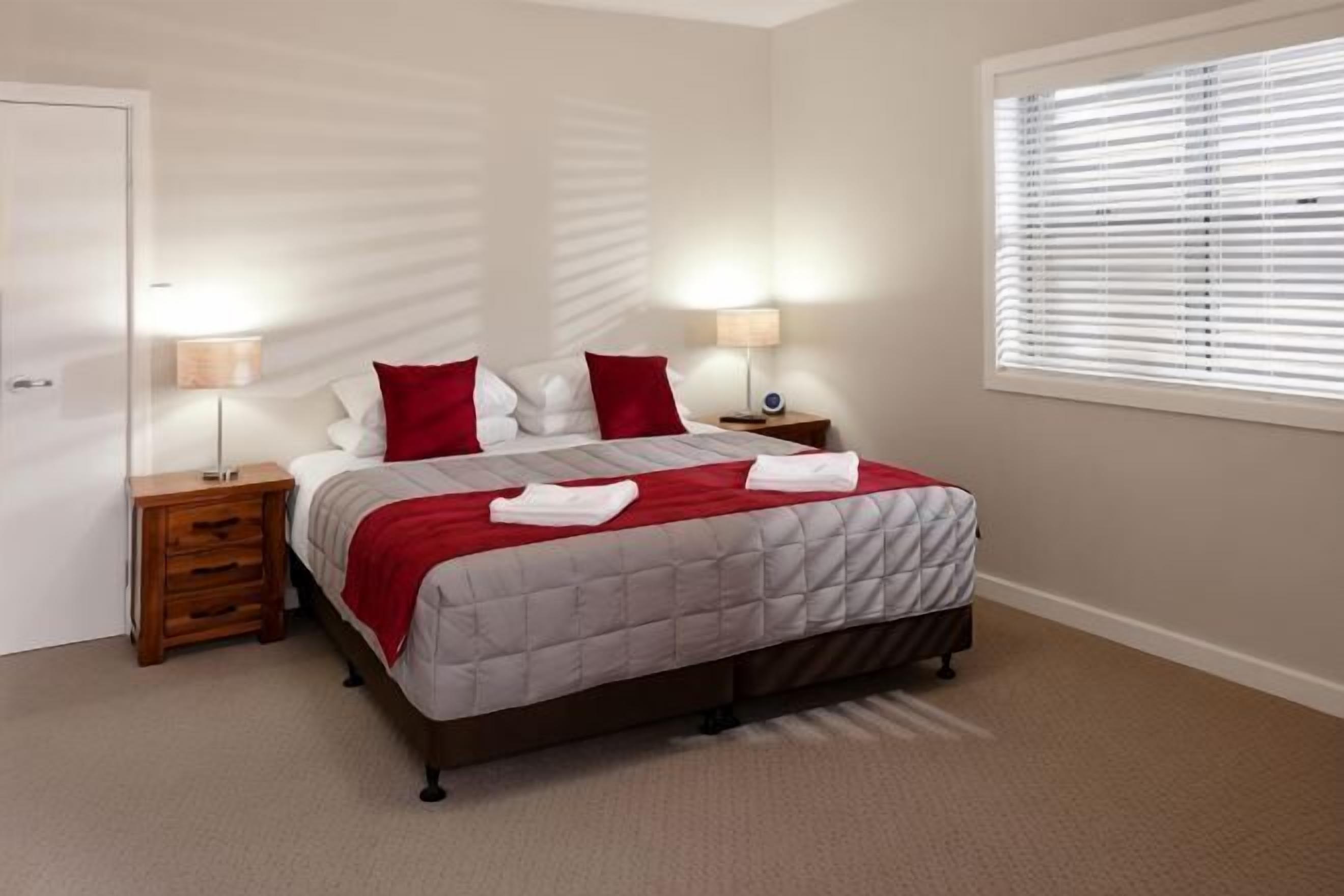 A bedroom with a bed, two bedside tables, a window with blinds, and a door.