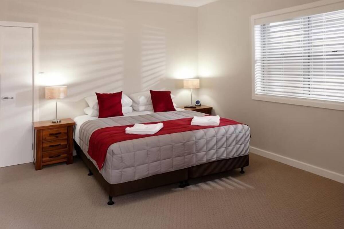 Um quarto com uma cama, duas mesas de cabeceira, uma janela com persianas e uma porta.