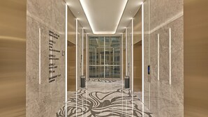 Un moderno vestíbulo de hotel con suelo de mármol, un ascensor de vidrio y un cartel direccional que indica los pisos y servicios del hotel.