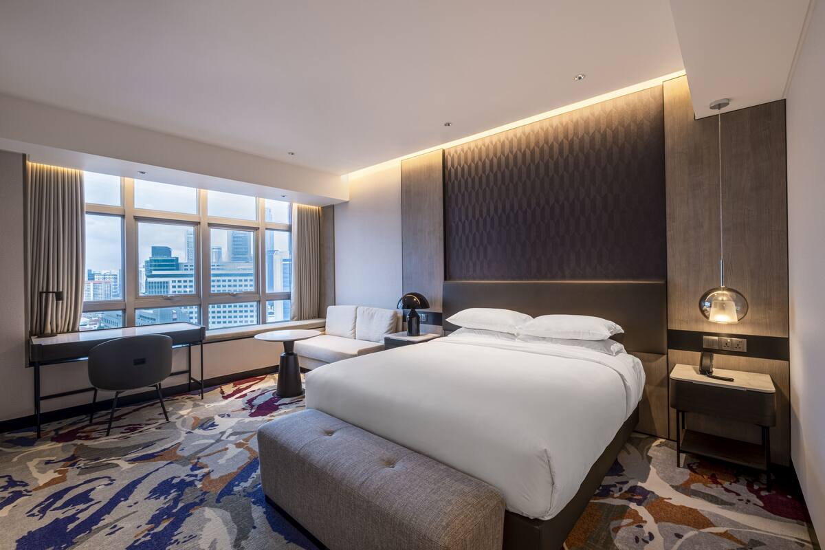 Quarto de hotel moderno com uma cama grande, uma escrivaninha e vista para a paisagem urbana através de amplas janelas.