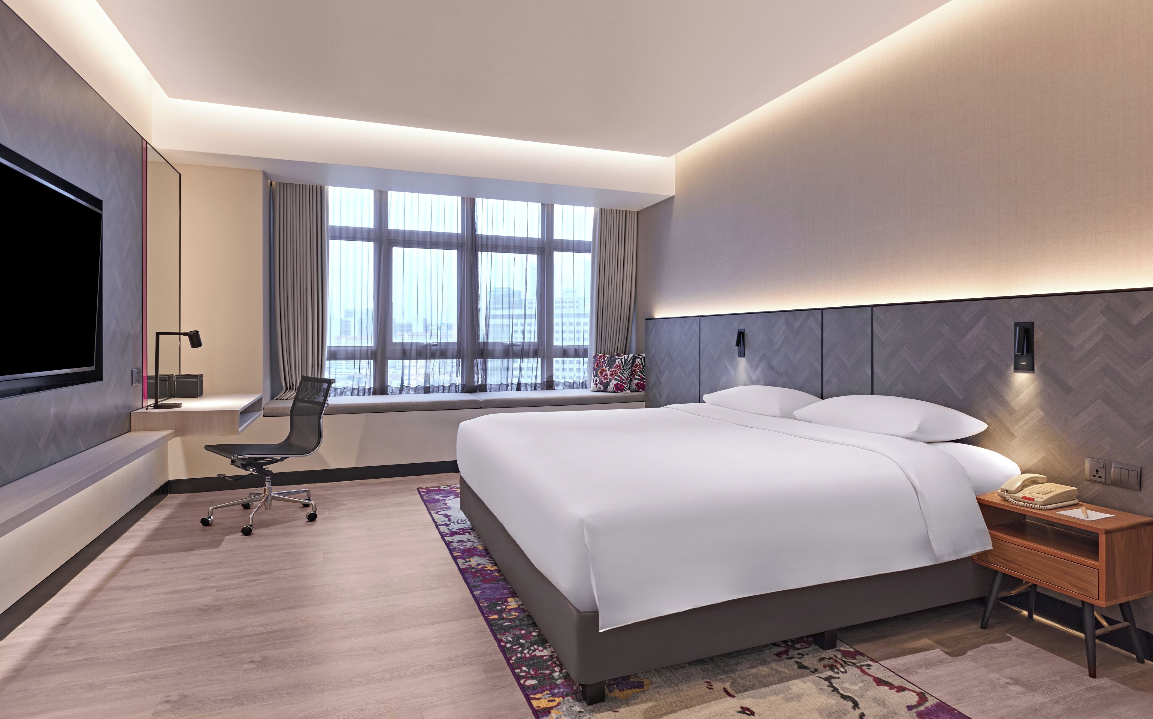 Una habitación de hotel moderna con una cama grande, un escritorio, una silla, un televisor y un ventanal con cortinas.