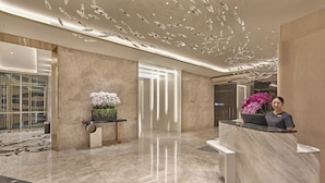 Un moderno vestíbulo de hotel con un mostrador de recepción de mármol, una planta en maceta y una lámpara decorativa en el techo.