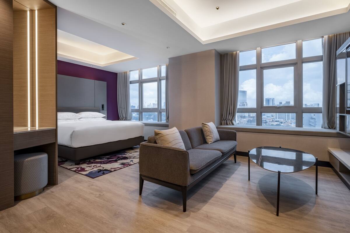 Quarto de hotel moderno com uma cama grande, um sofá, uma mesa de centro de vidro e vista para a cidade por amplas janelas.