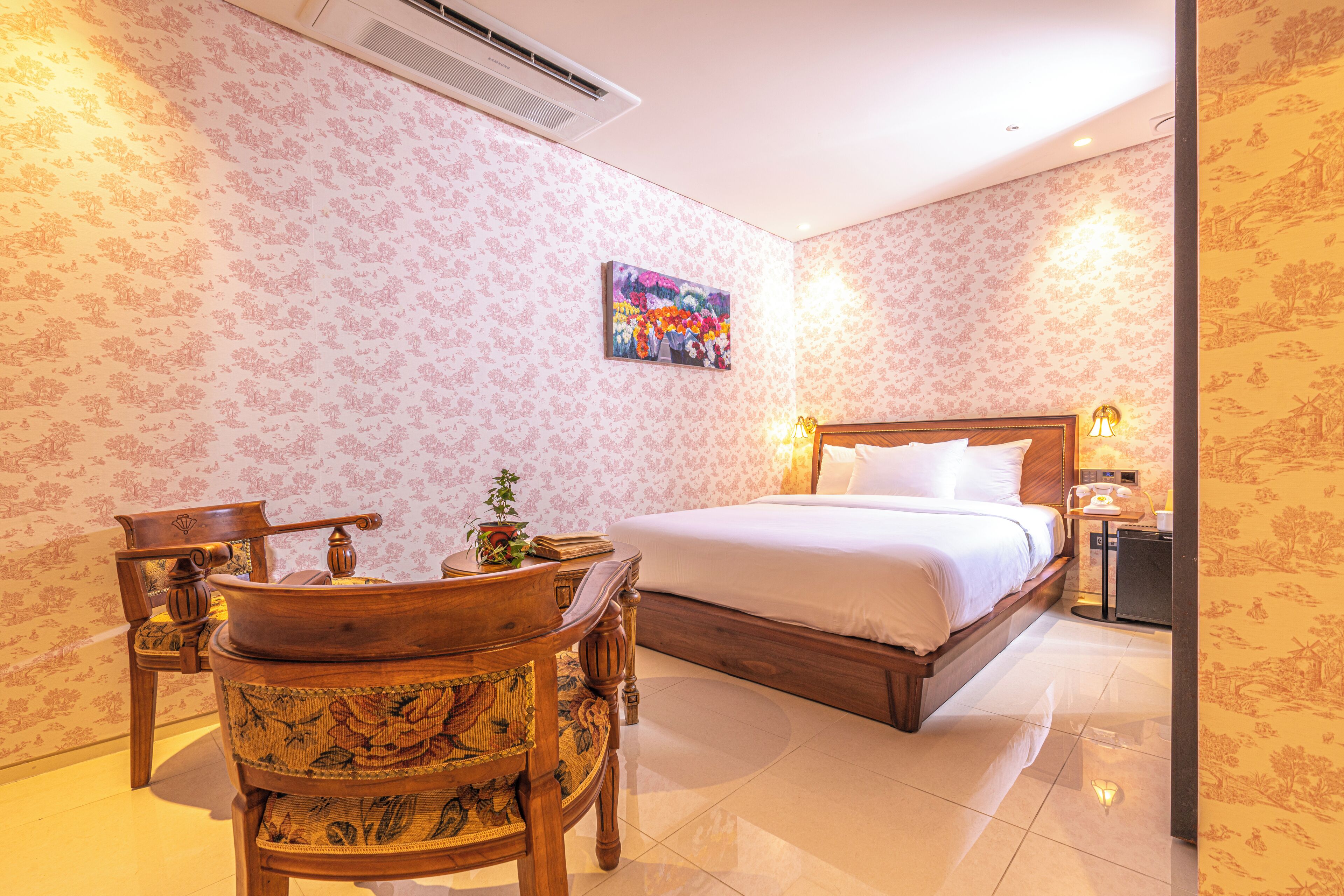 Quarto de hotel com uma cama, uma cadeira de madeira, uma mesinha e um quadro na parede.