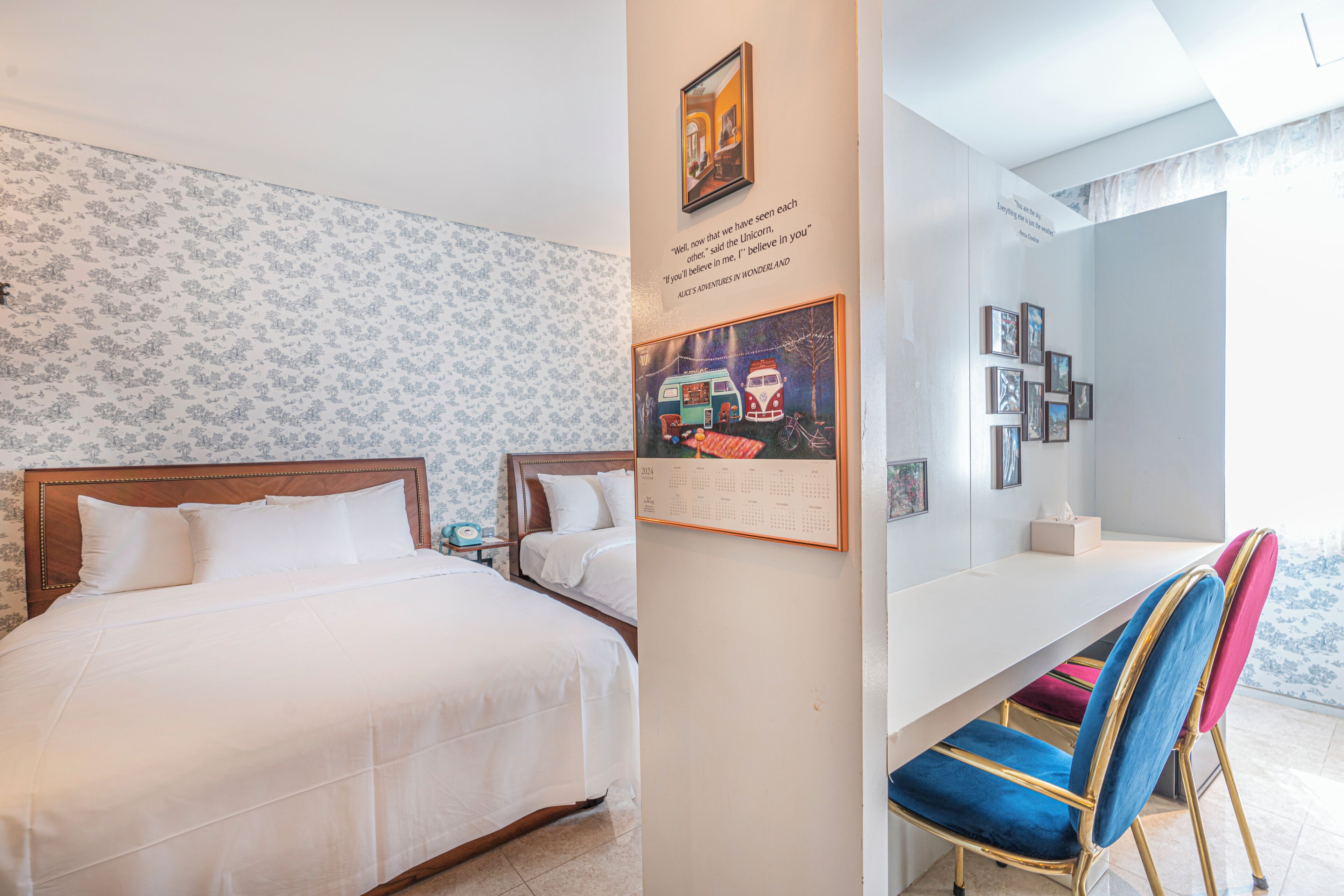 A hotel room with two beds, a desk, and a chair. There are framed pictures on the wall and a calendar.