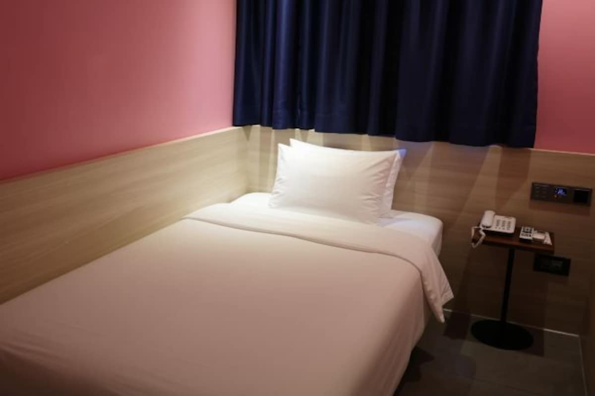 A neatly made bed with white linens in a room with pink walls and a dark curtain.