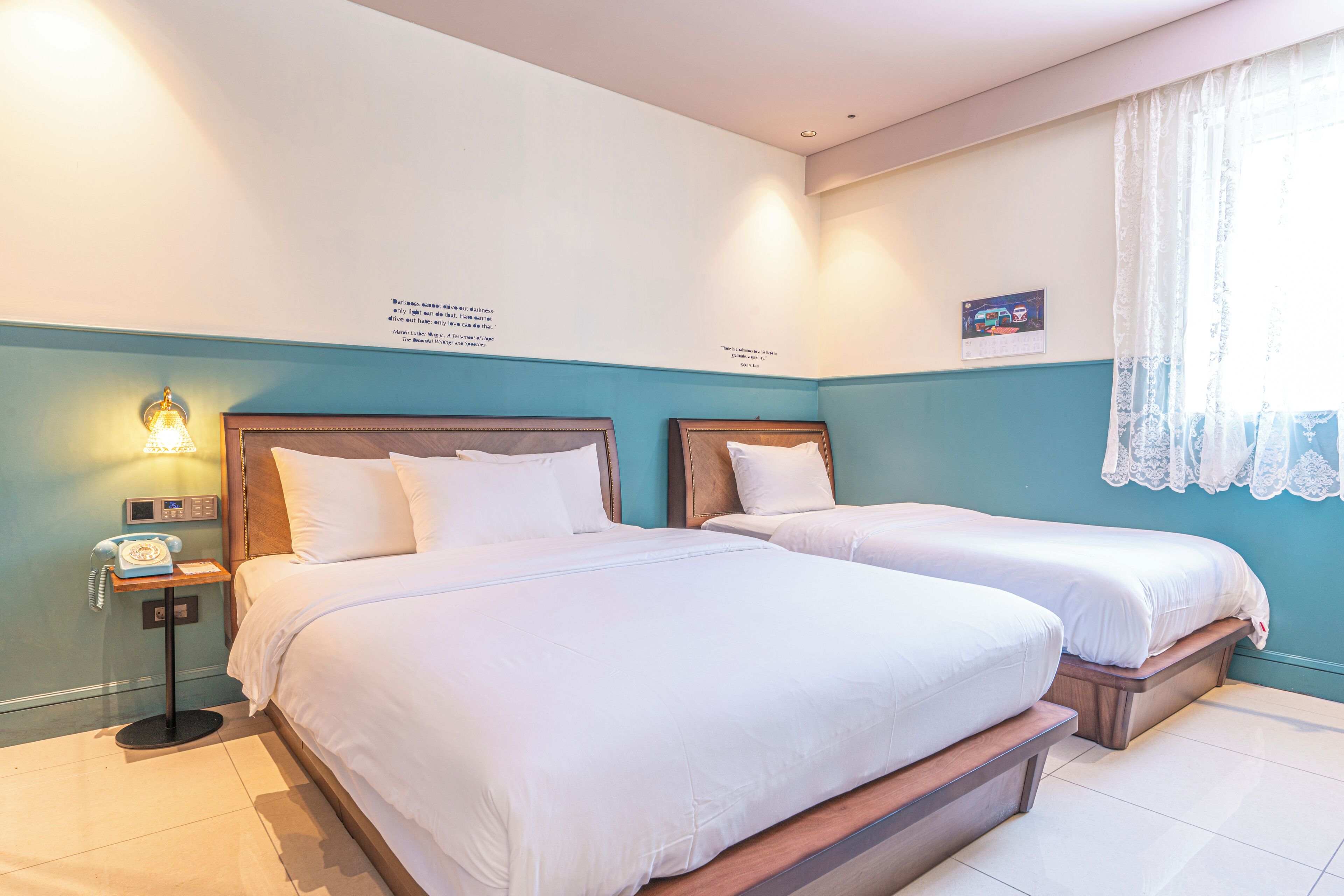 Ein Hotelzimmer mit zwei Betten, einem Nachttisch mit Lampe und einem Fenster mit Spitzenvorhängen.