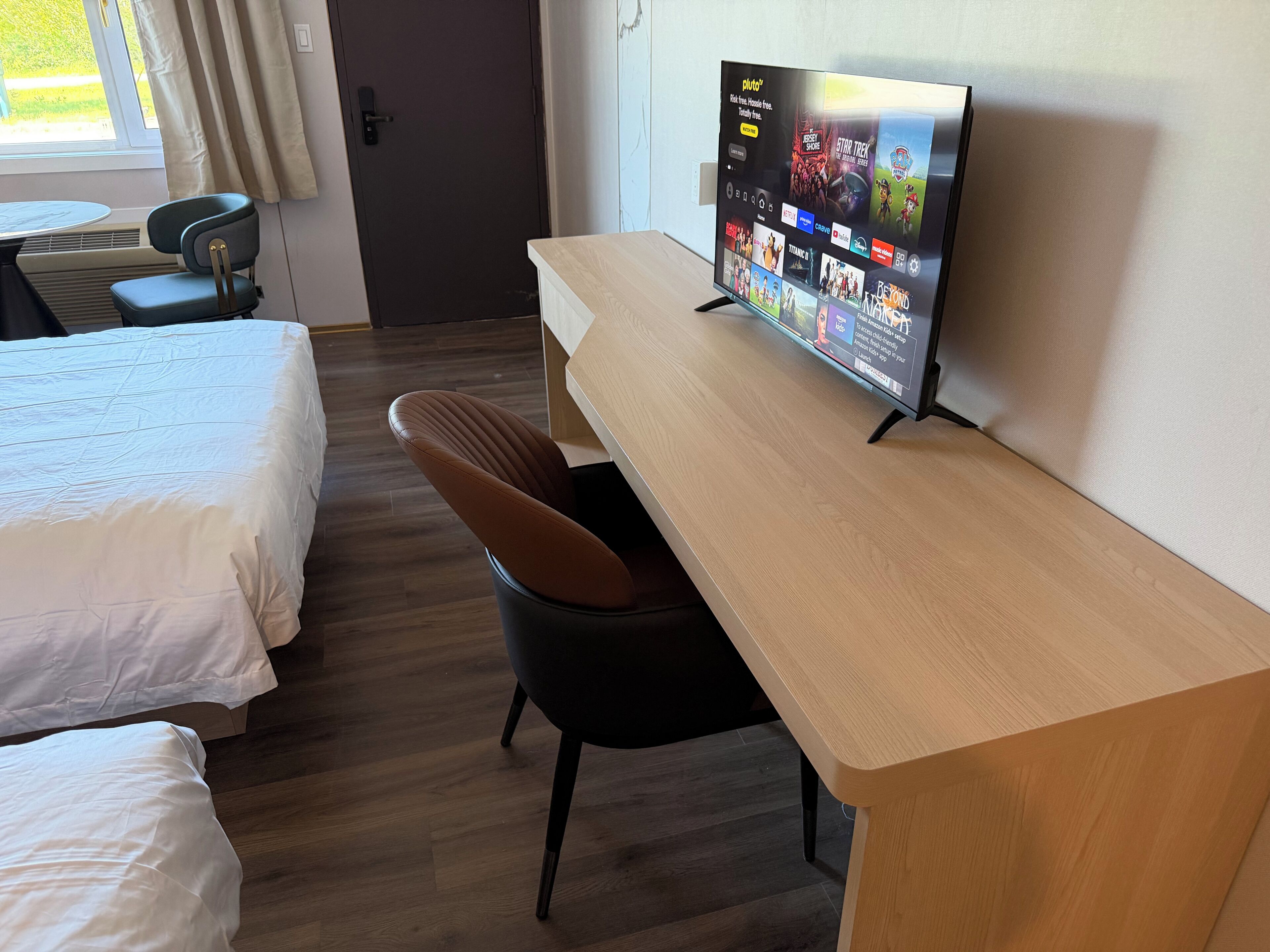 Quarto de hotel com cama, escrivaninha, cadeira, televisão exibindo a interface de um serviço de streaming e uma janela com cortinas.