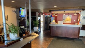 Un área de recepción con mostrador, plantas en macetas, una pantalla digital y un letrero de bar.