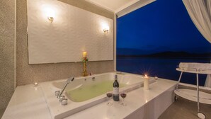 Um banheiro moderno com uma banheira grande, um espelho e vista para o mar ao entardecer.