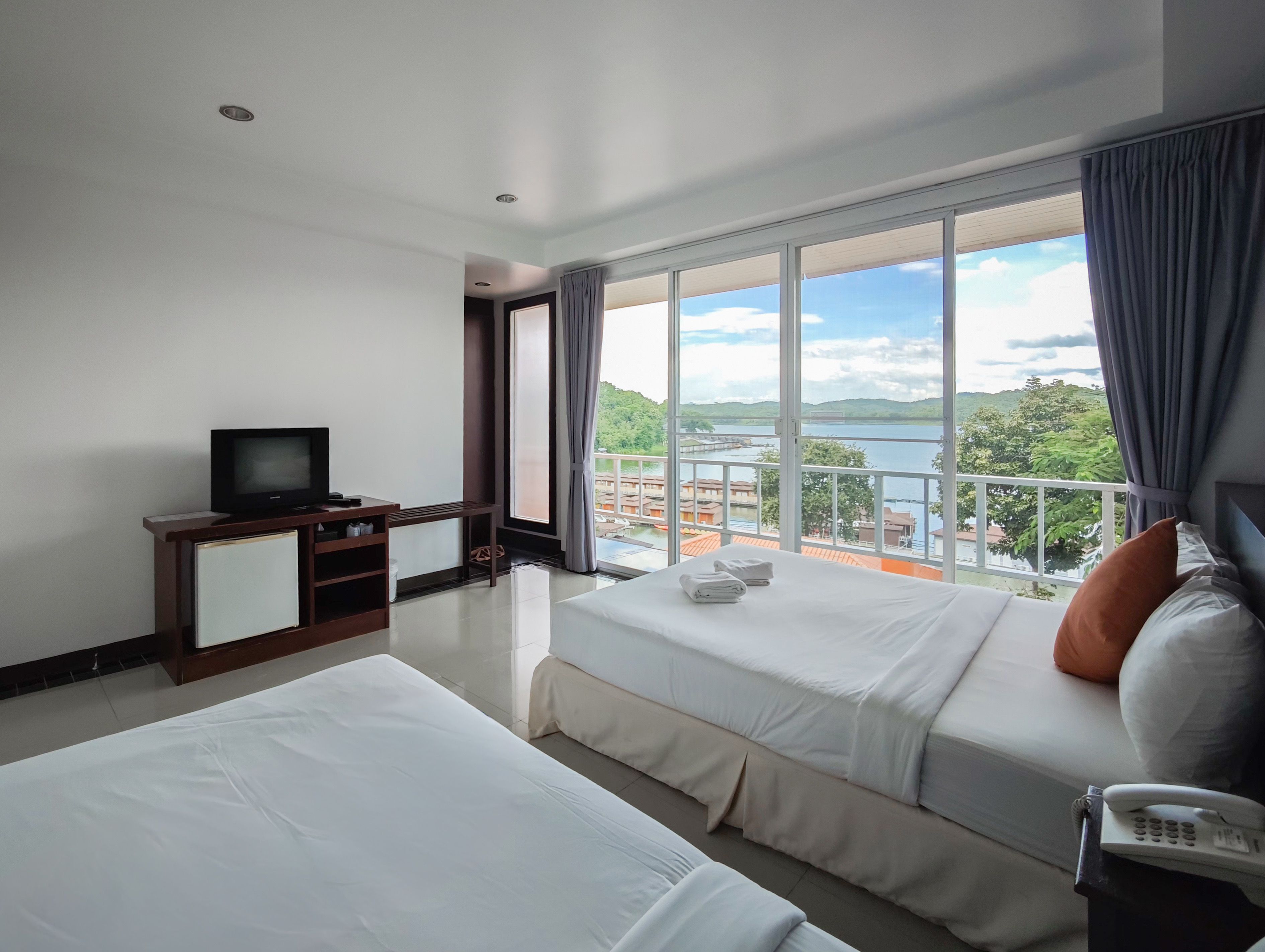 A hotel room with two beds, a TV, and a balcony with a view of the water and buildings.