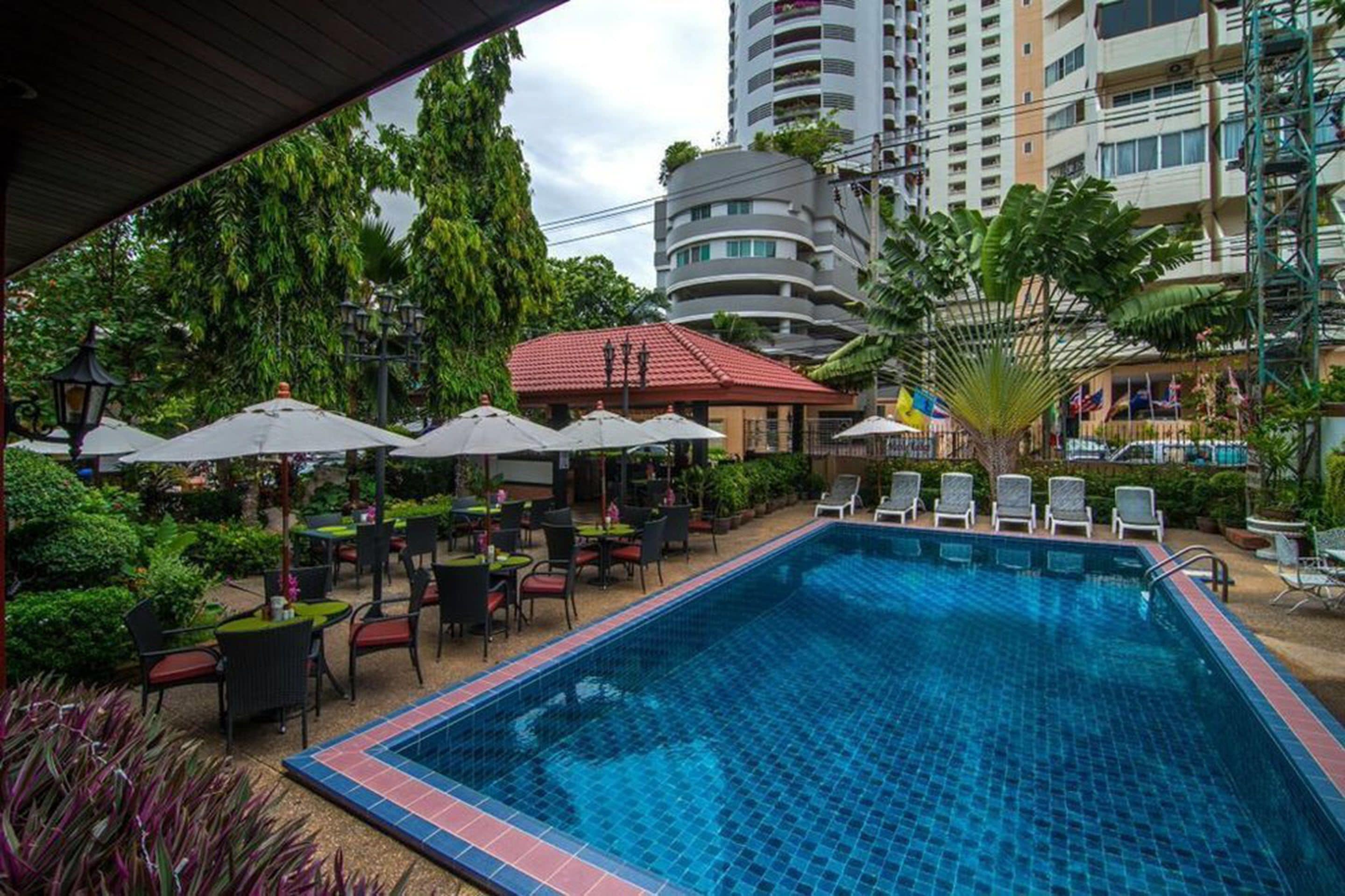A swimming pool with lounge chairs and umbrellas, surrounded by greenery and a building in the background.