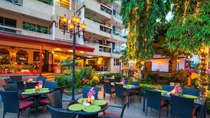 An outdoor dining area with wicker chairs and round tables, surrounded by lush greenery and decorative lights.