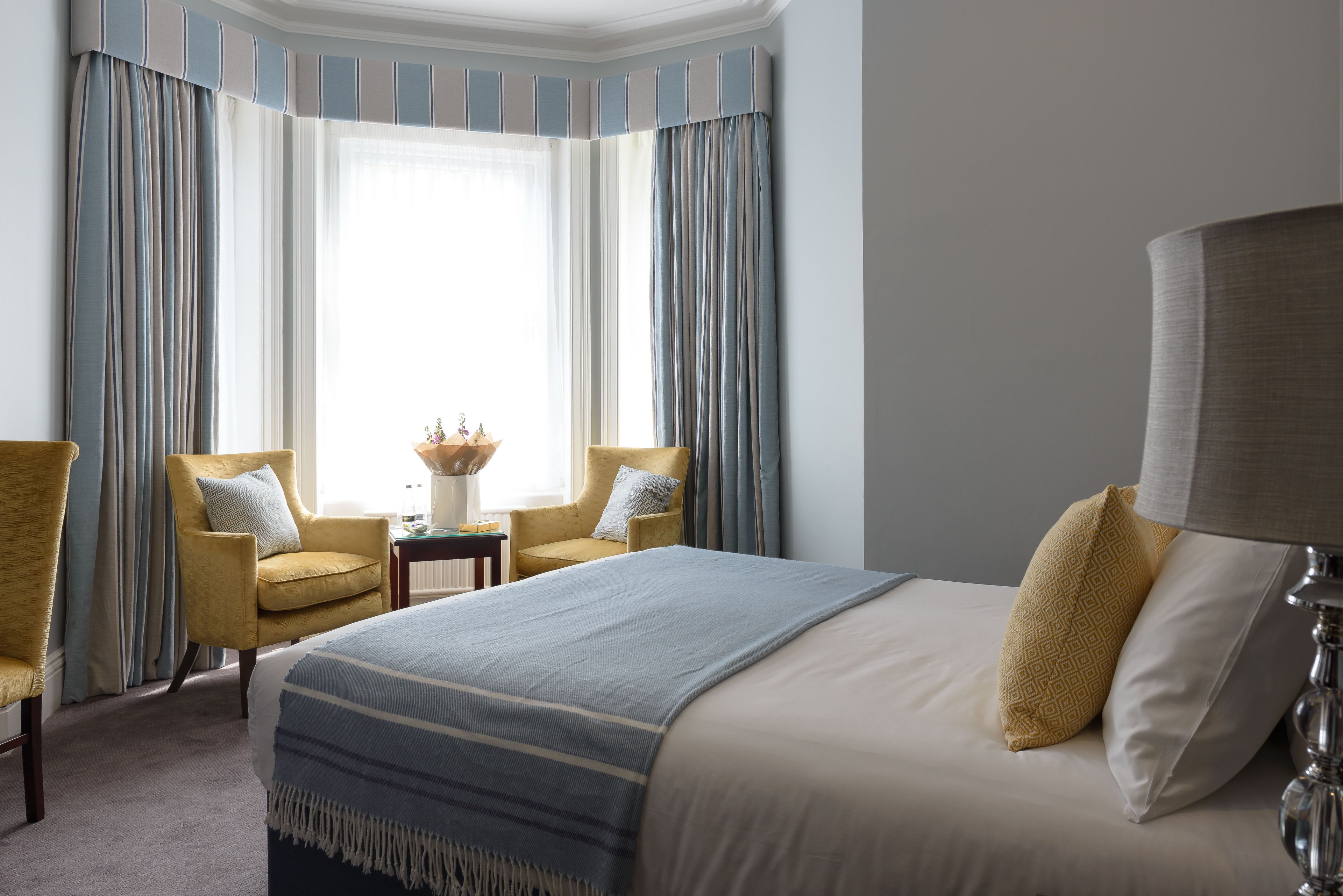 A hotel room with a large bed, two armchairs, a small table with a vase, and a window with curtains.