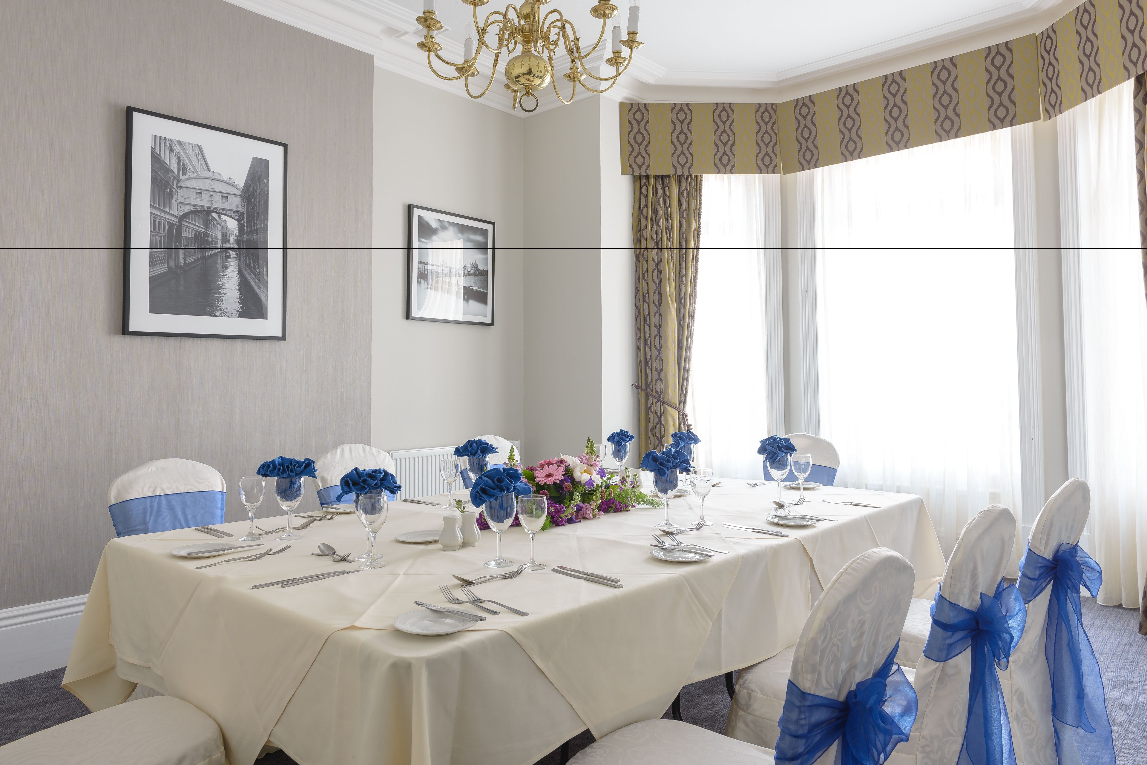 A dining room with a long table set for a meal, chairs with blue sashes, and framed pictures on the walls.