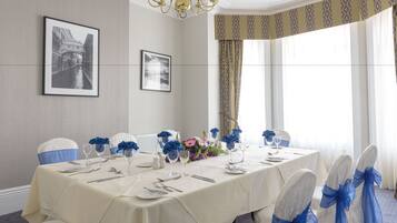 A dining room with a long table set for a meal, chairs with blue sashes, and framed pictures on the walls.