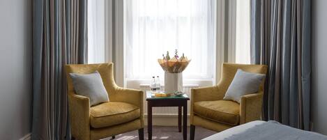 A hotel room with two yellow armchairs, a small table with a vase, and a bed.