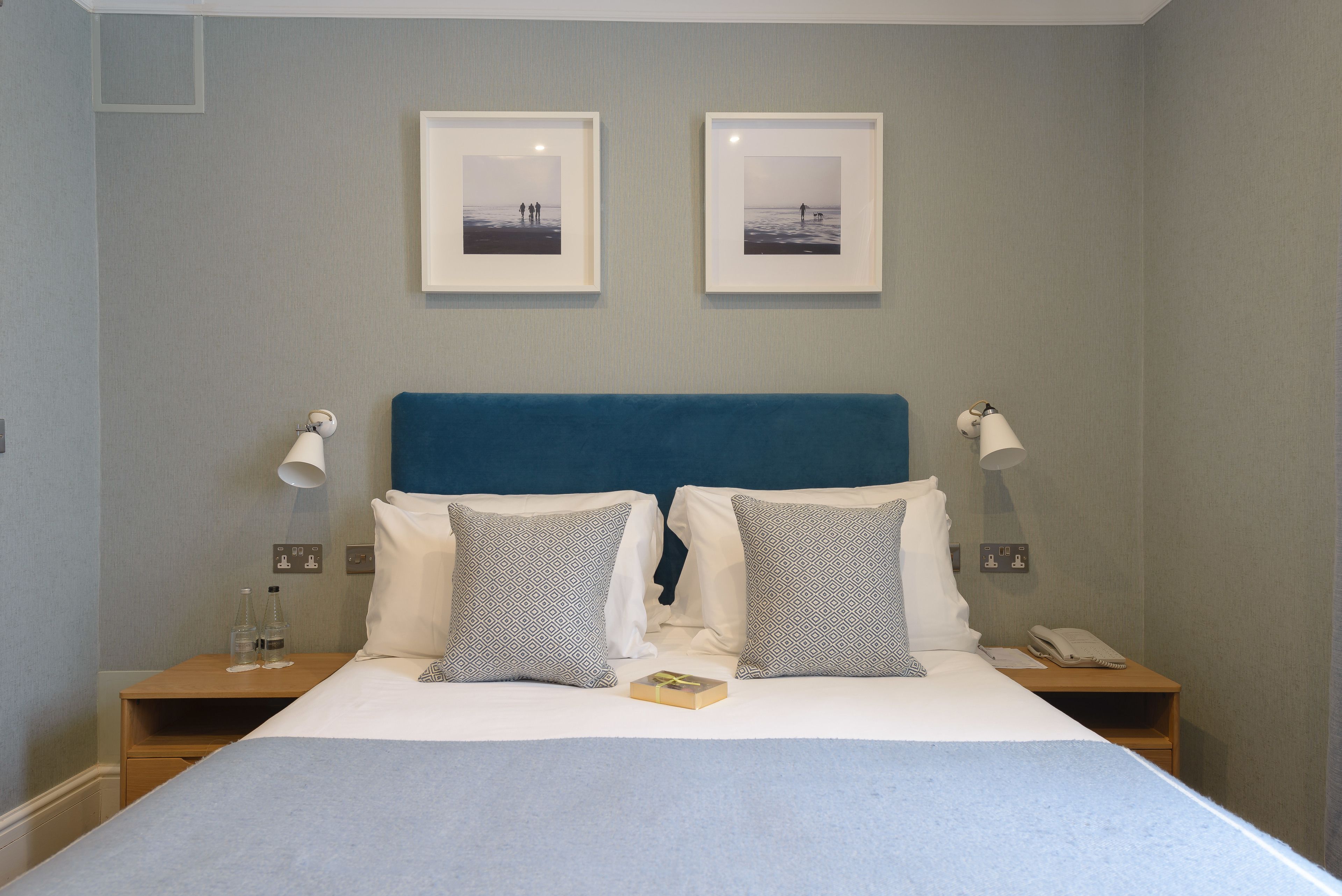 A neatly made bed with a blue headboard, two bedside tables with lamps, and two framed pictures on the wall.