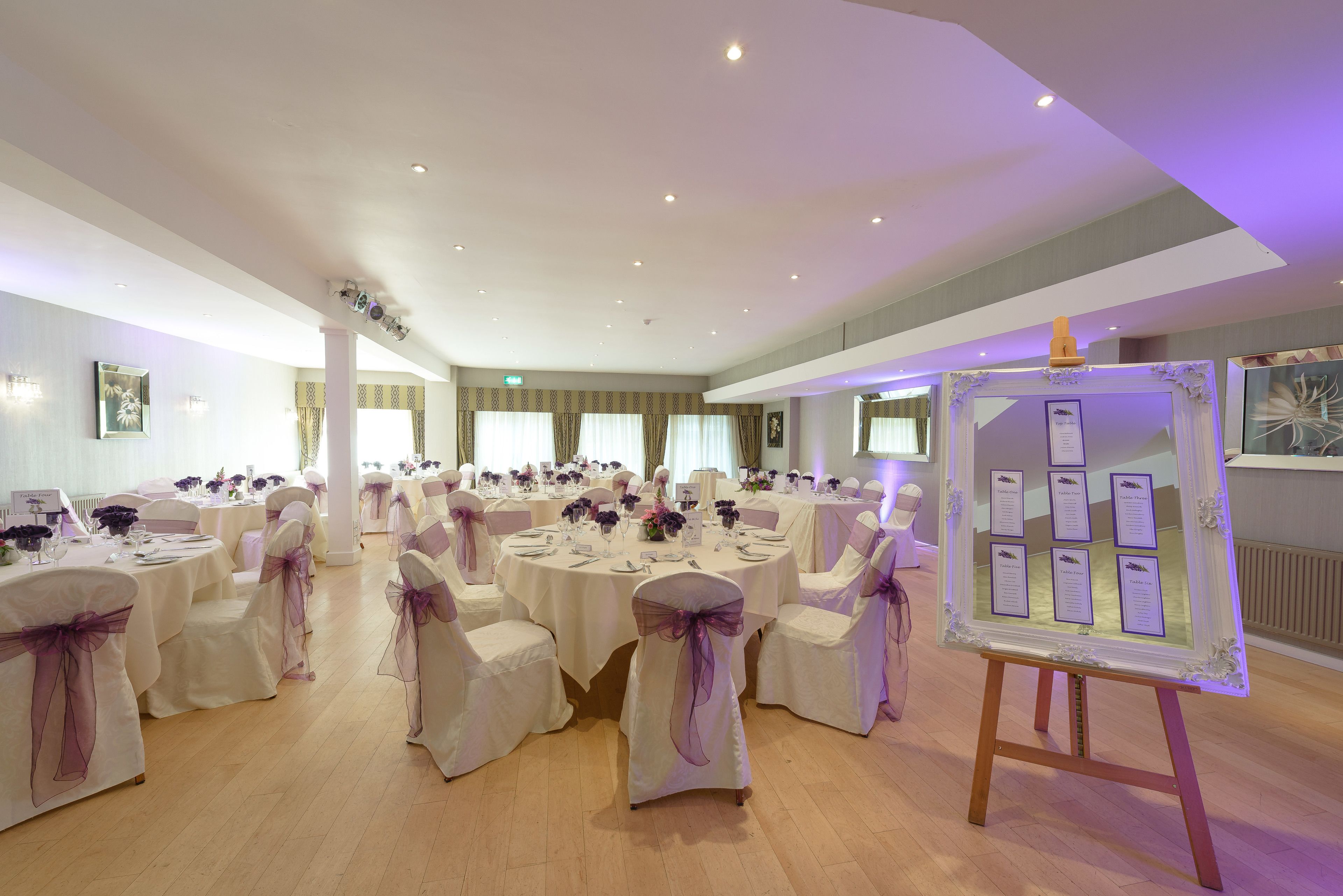 A banquet hall with round tables, white chairs with purple sashes, and a framed display board.