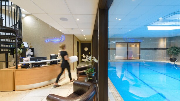 A hotel lobby with a reception desk, a pool area visible through the glass, and a staircase.
