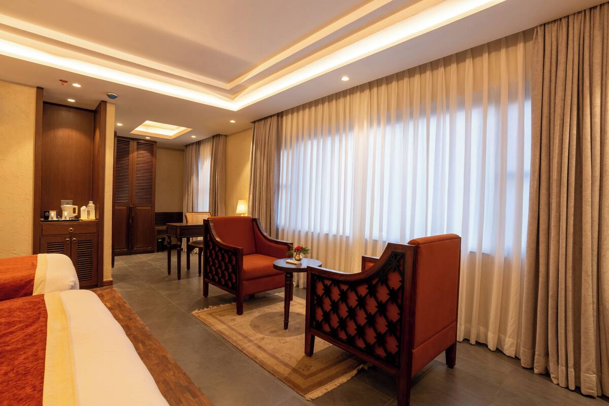 Habitación de hotel con una cama, dos sillones, una mesita, un escritorio, una lámpara y un amplio ventanal con cortinas.