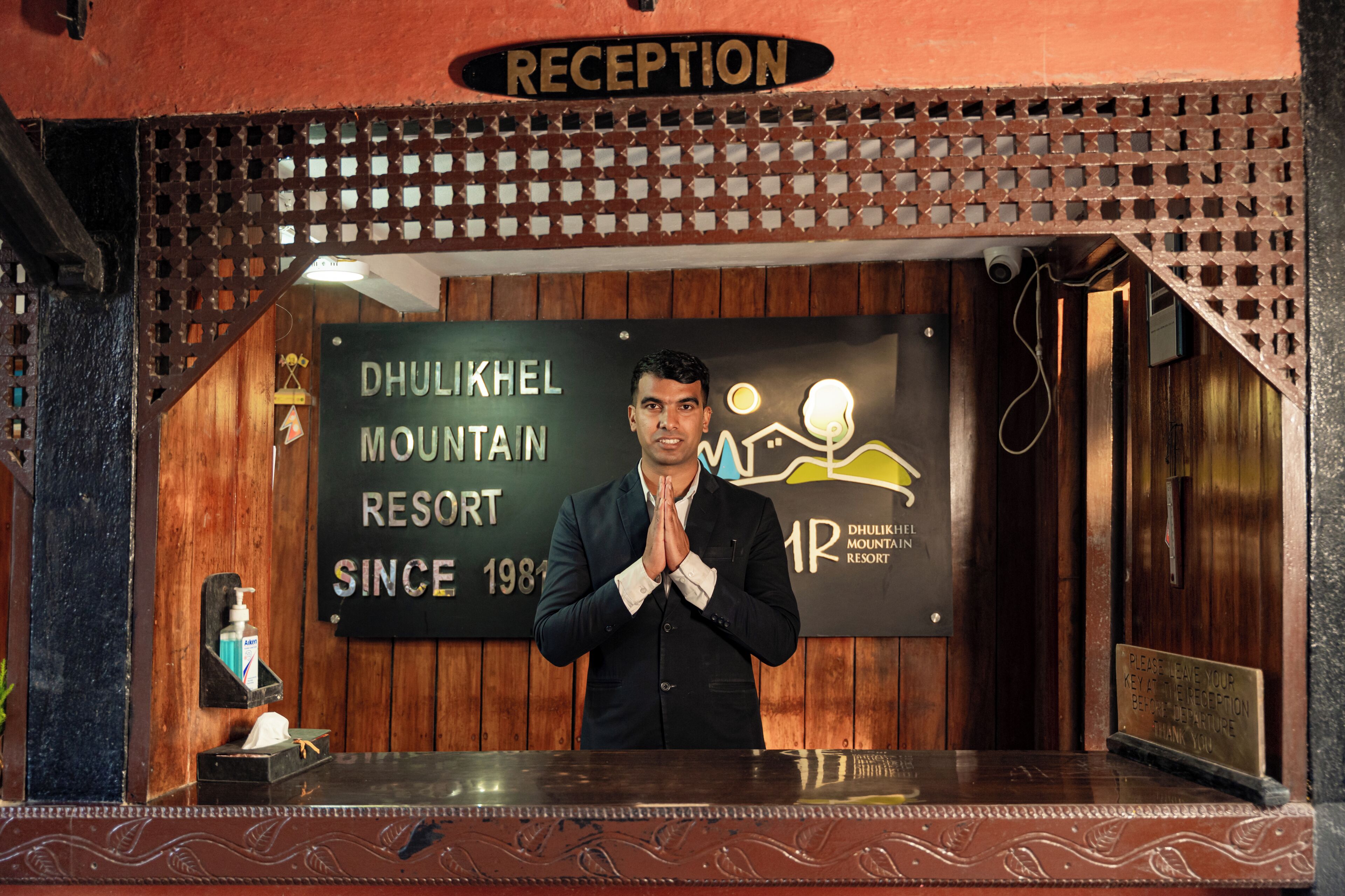 Modtagelsesdisken på Dhulikhel Mountain Resort, indrettet med træ, et skilt og en mand, der står med foldede hænder.