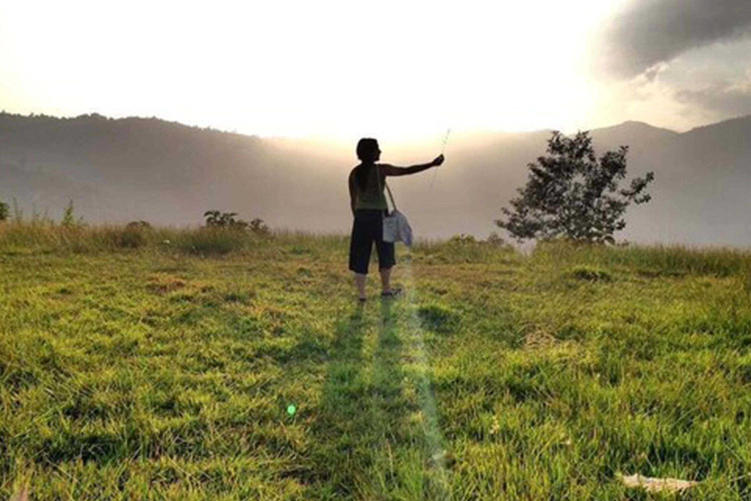 A person standing in a grassy field with a tree and mountains in the background.